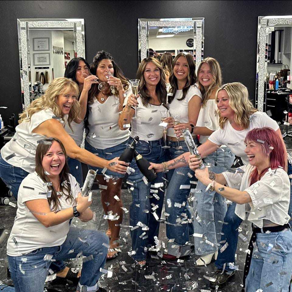 Group of women in white shirts and jeans celebrating with champagne and confetti in a salon.