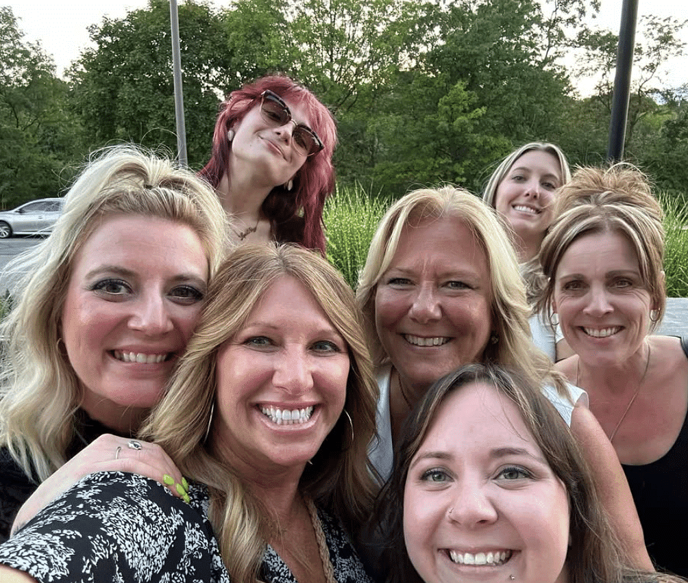 Group of smiling women outdoors with trees in the background.